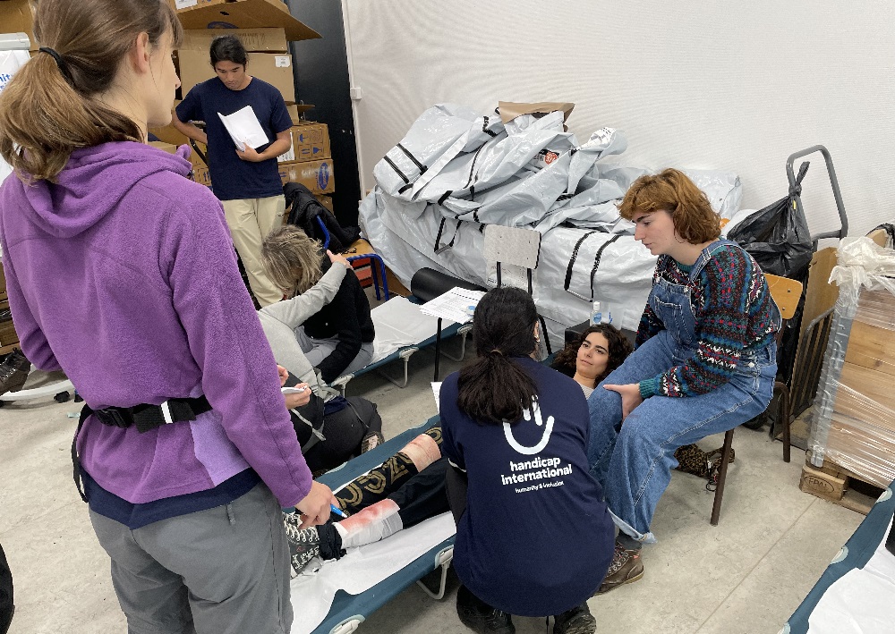 Exercice de simulation : personnels de HI au chevet de blessés allongés sur des brancards au sol dans une salle qui sert d'hôpital et dépôt logistique.