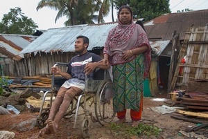 Devant des maisons détruites par des inondations, un homme en fauteuil roulant et une femme debout.