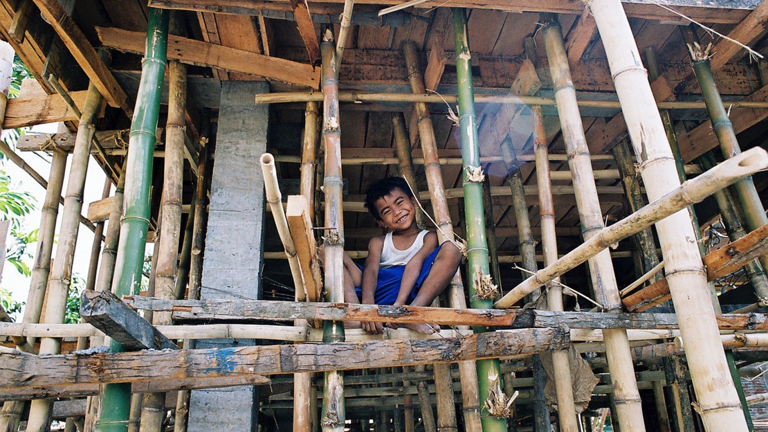 Un petit garçon est accroupi avec un grand sourire au milieu de son habitation faite de bambou.