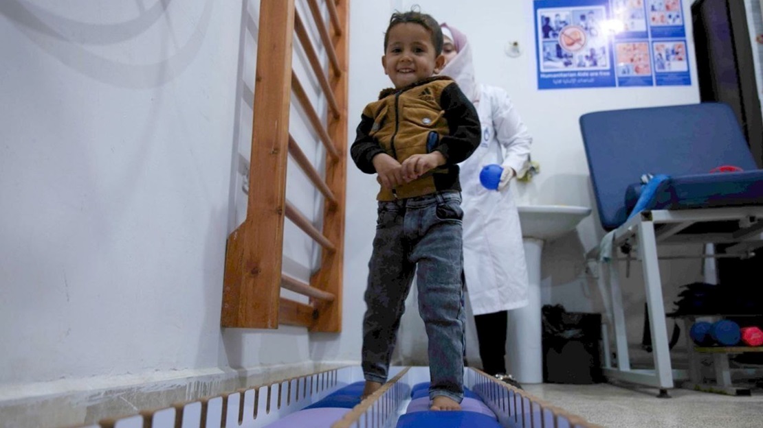A young boy walks on a foam mat in a rehabilitation room while a woman stands beside him.; }}