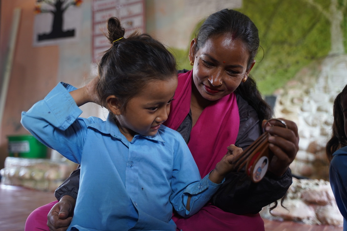 Aakanksha, 6 , and her teacher, Nita, using the communication keyring provided by HI to help her make herself understood better in class. A little girl is looking at her communication keyring, held out by her teacher. The two of them are smiling at each other.; }}