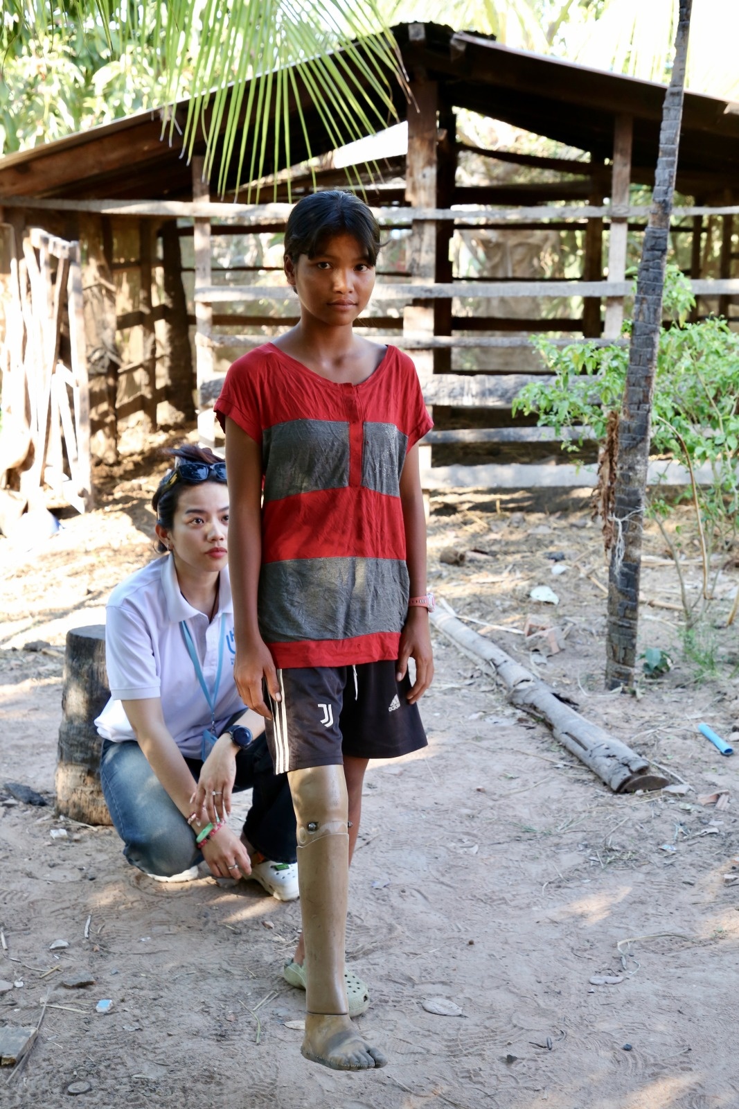 A young girl fitted with a prosthetic is about to walk as Rakseymutta just adjusted her assistive device and is sitted behing her