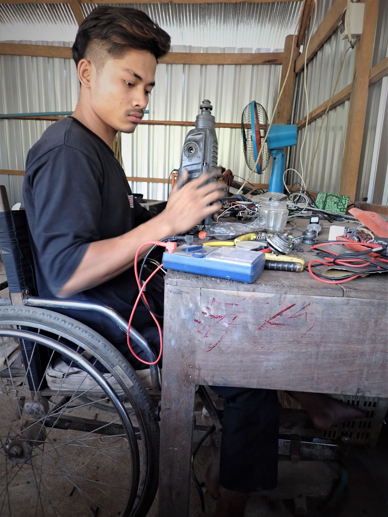 Heang Tienglin possède son atelier de réparation électronique à proximité de son domicile. Il répare une perceuse, assis dans son fauteuil roulant contre son établi. 
