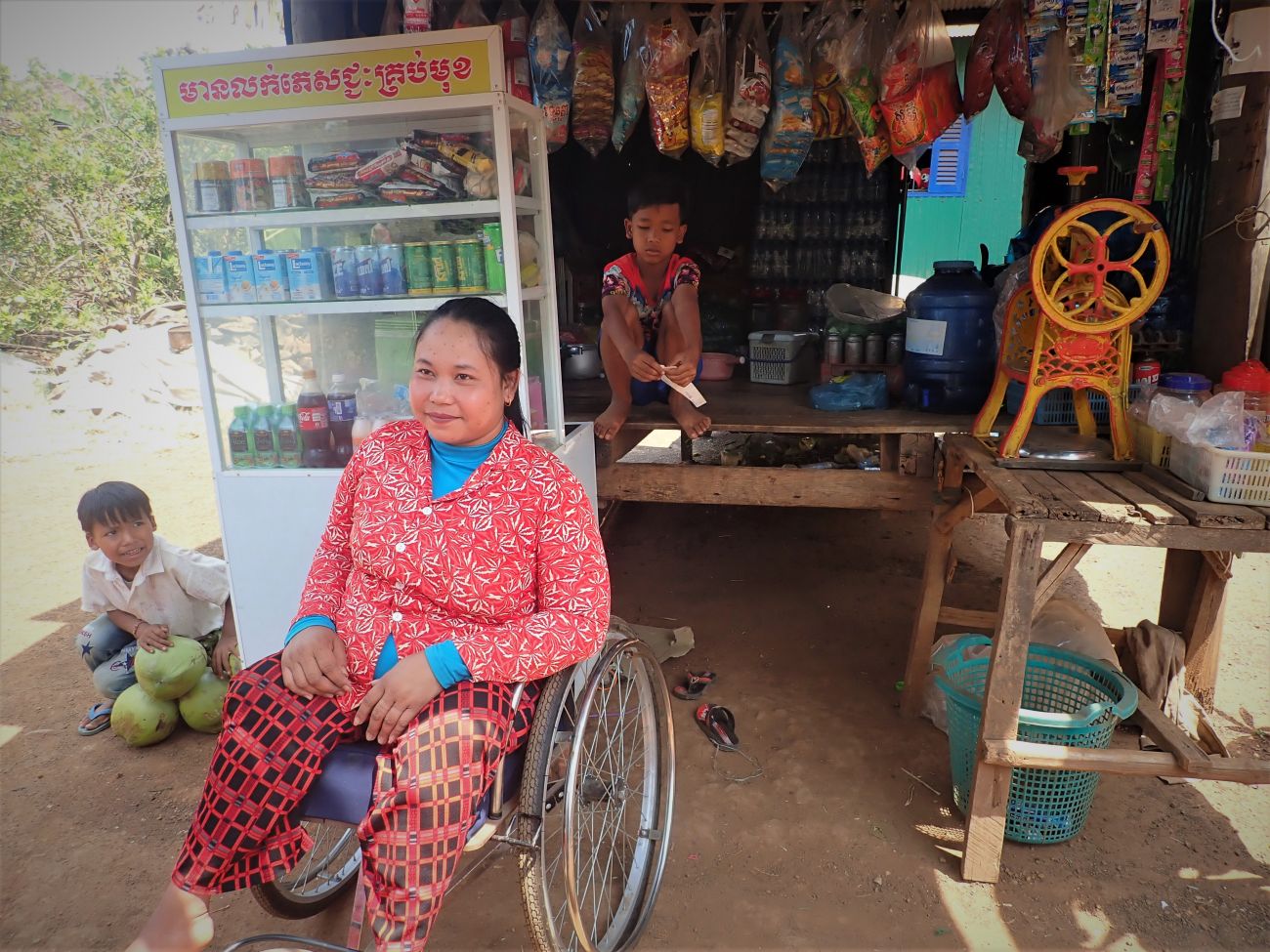 Min Sokly vend des boissons et de la nourriture dans sa petite échoppe, ouverte grâce au soutien de HI. La jeune femme se trouve devant en fauteuil roulant entourée de deux enfants.