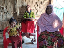 Une mère de famille et ses deux filles posent pour la photo, au premier plan à gauche une petite fille avec une couette et un haut jaune, sa maman à droite porte une robe avec des lys et un voile rose, au fond une adolescente, sa fille aînée en fauteuil roulant. Ells sont à l'intérieur de leur abri.