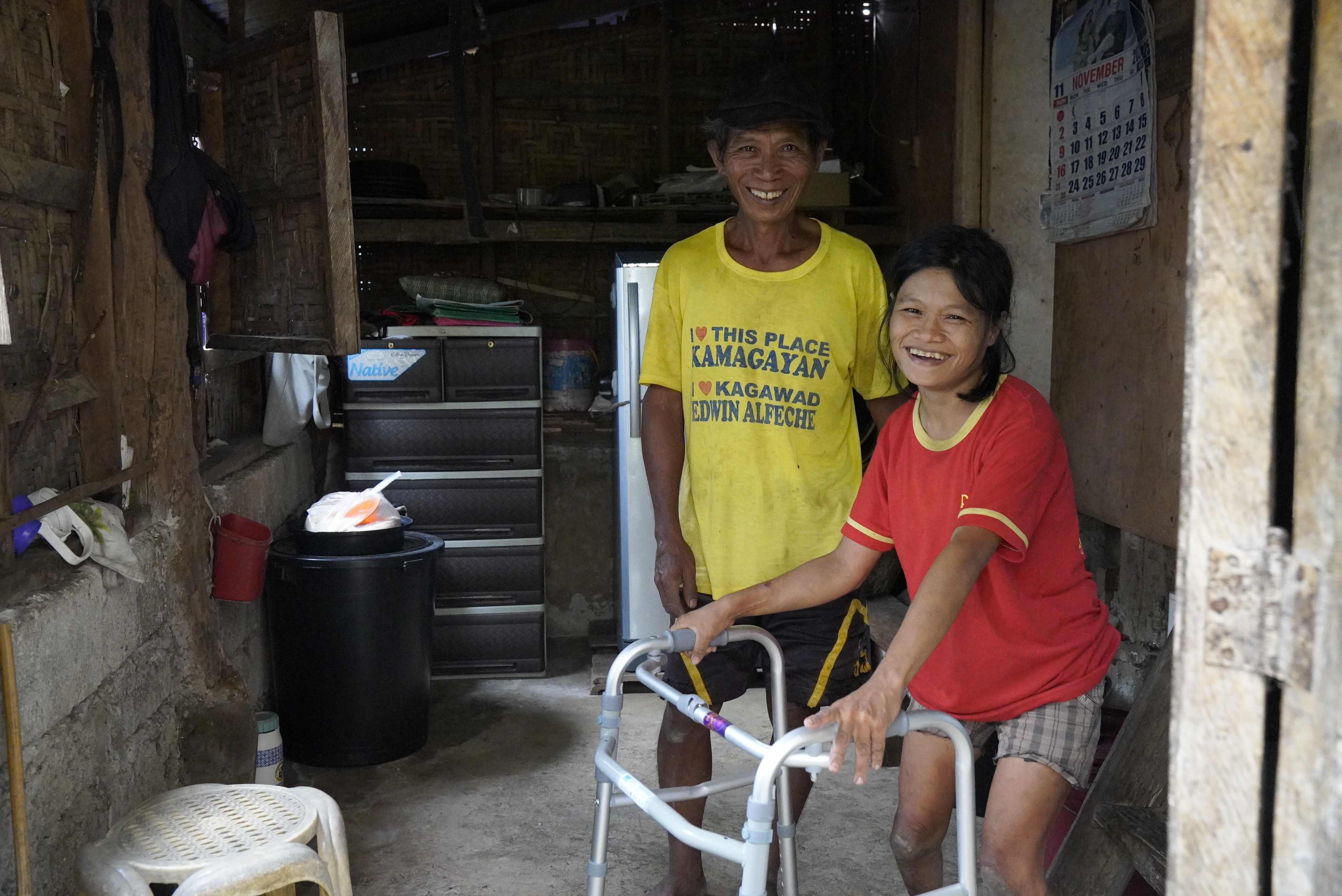 Een vrouw met een rollator lacht naar de camera. Een andere man staat naast haar.