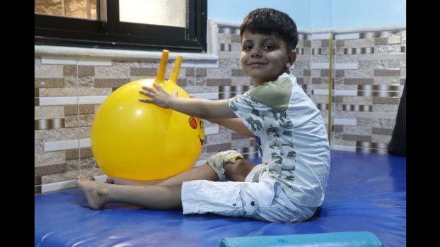 Ayham est assis sur un tapis et utilise un ballon pour l'aider dans sa rééducation.; }}