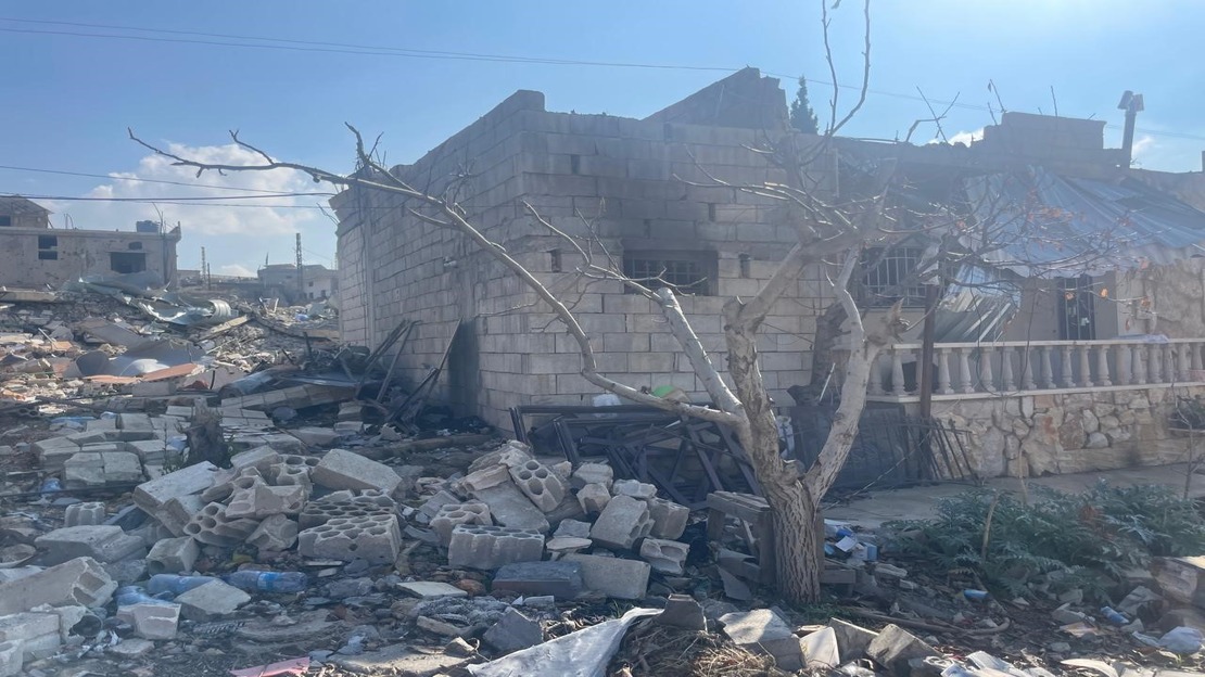 The photo shows a partially destroyed brick building, whose roof and some walls appear to have collapsed. In the foreground, a leafless tree stands amid a large pile of rubble: broken concrete blocks, pieces of wood, metal fragments, and various scattered objects. To the right, another structure, built of stone and covered with damaged sheet metal, also appears to have been damaged. The ground is completely littered with rubble, and in the background, other damaged buildings can be seen under a clear sky.  ; }}