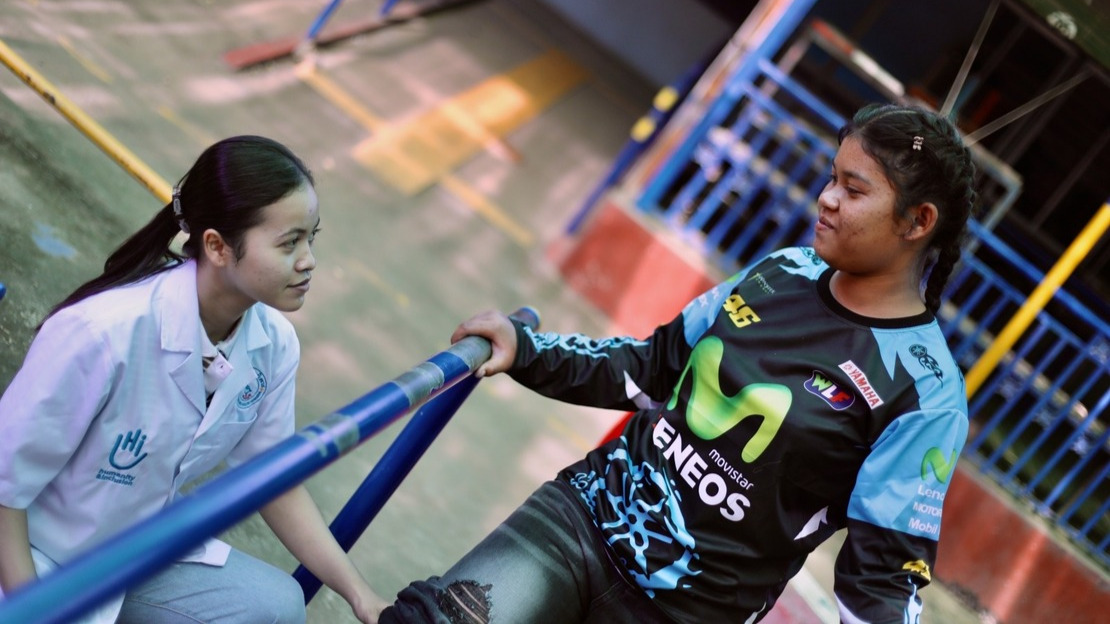 A young girl stands between two physiotherapy exercise bars and looks down at a lady crouching beside her wearing a HI-branded smock.