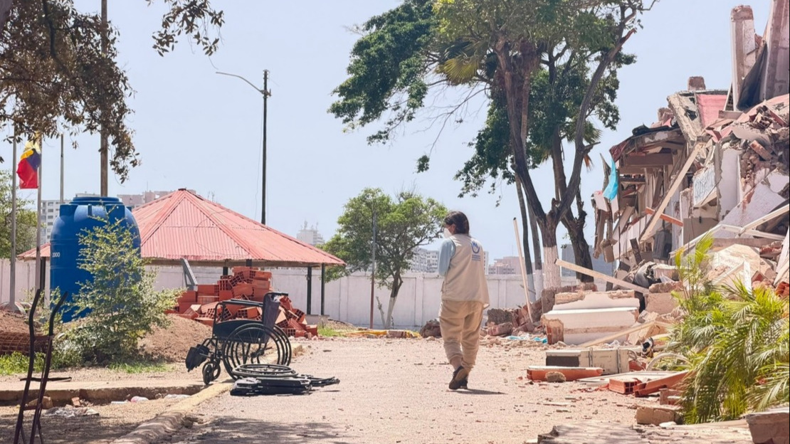 Une femme se tient de dos dans une rue terreuse entourée de décombre de bâtiments. Elle regarde sur la gauche deux fauteuils roulants abandonnés.