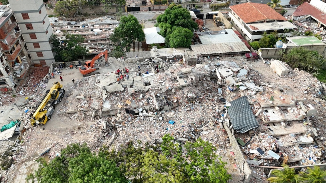 A drone view of a group of buildings that have been completely destroyed. Service vehicles and people can be seen amidst the rubble.; }}