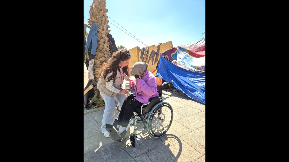 Une personne est assise dans un fauteuil roulant à l’extérieur et porte une veste violette et un foulard sur la tête. Le fauteuil roulant est positionné sur un sol pavé.  Une personne debout se penche vers la personne assise dans le fauteuil roulant et lui tient le coude. En arrière plan, on voit une tente bleue, des vêtements suspendus à des cordes et un palmier, sous un ciel dégagé.