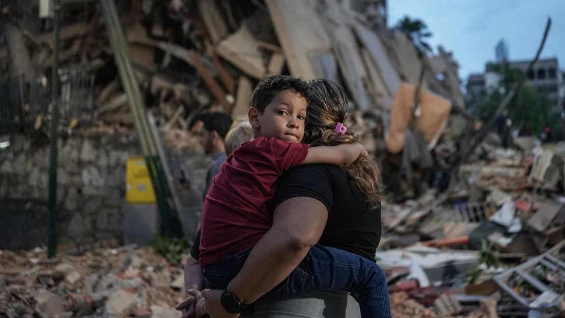 Au premier plan, une femme de dos porte dans ses bras un enfant qui regarde fixement la caméra. Derrière eux, on voit des bâtiments effondrés.