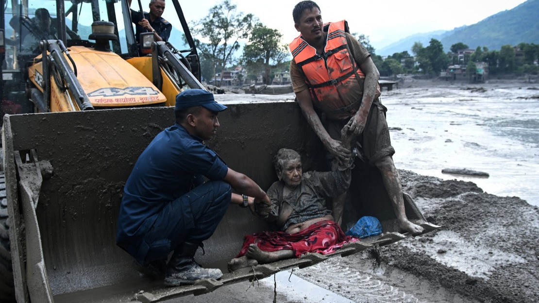 Two men are supporting a disabled woman who is half-reclining in the bucket of a backhoe loader. They are all covered in mud, and the landscape around them is flooded.; }}