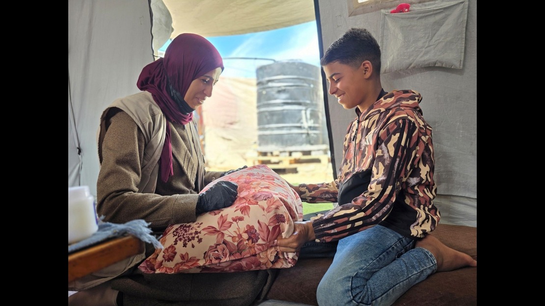 Inside a canvas shelter, two people are sitting facing each other on cushions. An adult is wearing layered clothing and a scarf covering their hair, and is holding a floral-patterned cushion on their lap. Across from them, a younger person, kneeling on a rug, wears a patterned hoodie and jeans, and is also holding the cushion with their hands. In the background, an opening reveals a blue sky and hanging fabrics. Natural light illuminates the scene through the opening. ; }}