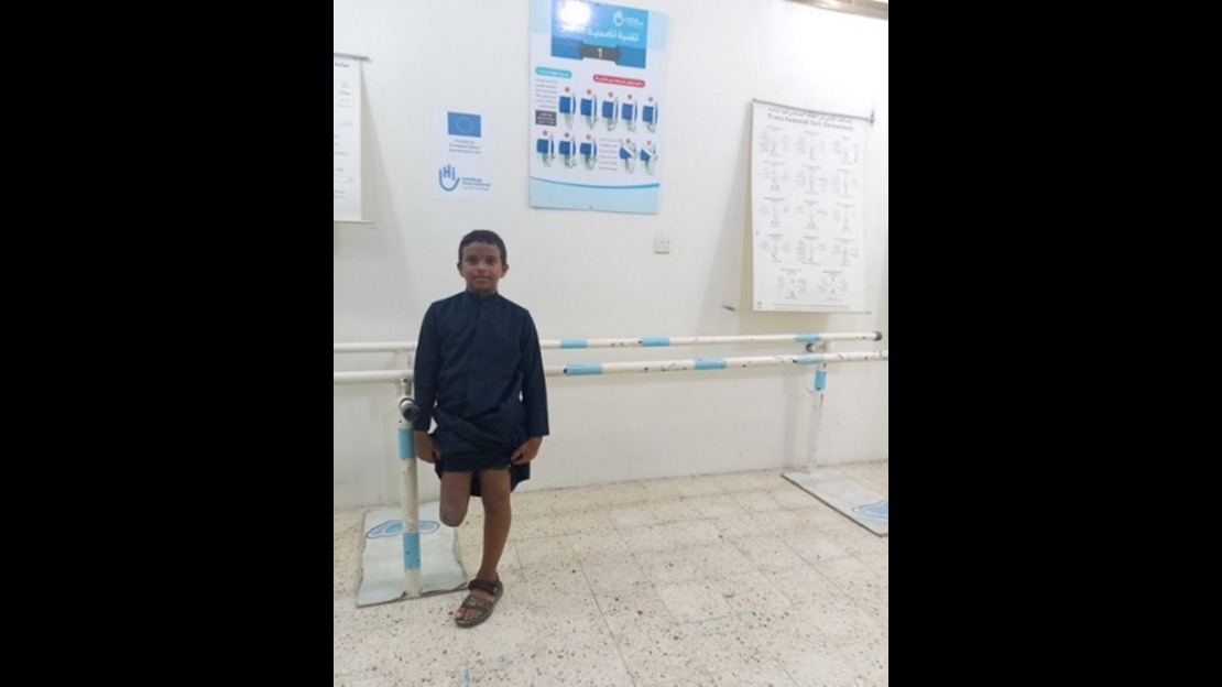 A young boy is standing in a physical therapy room, leaning slightly on white and blue parallel bars anchored to the floor, which are used for walking practice. The room is brightly lit, with medical and educational posters hanging on the wall in the background.; }}