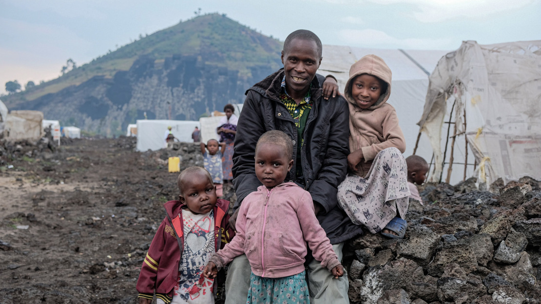 A smiling man is surrounded by three children. They are sitting on a low black stone wall. Behind them, rows of tents can be seen, and in the distance, a green hill.; }}