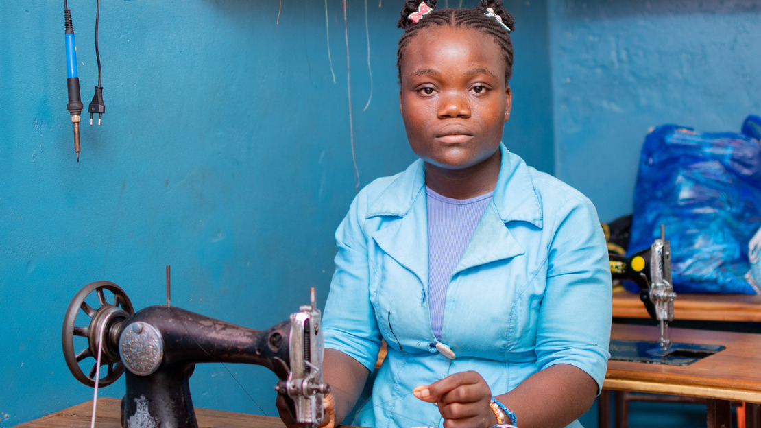 Portrait de Belvine assise face à une table et qui regarde la caméra. Devant elle est posée une machine à coudre ainsi qu'un morceau de tissu. Elle tire un fil passé dans la machine.; }}