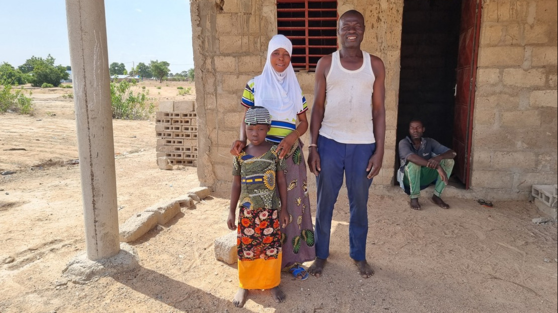 A man, a teenager and a little girl are standing in front of a brick house and smiling.; }}