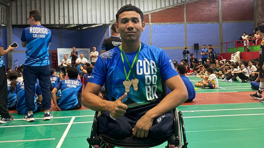 Un jeune homme en fauteuil porte un maillot sportif et a deux médailles passées autour du cou. Derrière lui, on devine un stade couvert.; }}