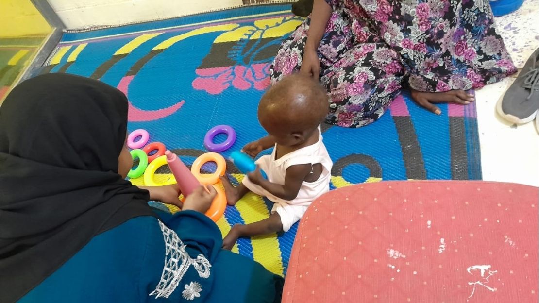 A child is sitting on a mat, holding a toy handed to him by a woman; }}