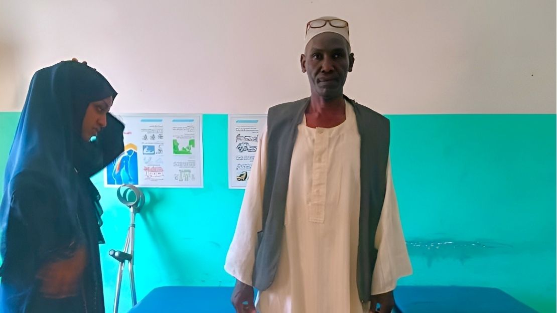 A man is standing and looking at the camera. In the background, a few crutches are leaning against the wall.; }}