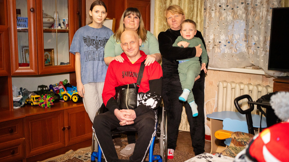 Un homme est assis dans un fauteuil roulant dans un appartement avec autour de lui sa famille.; }}
