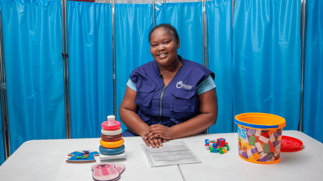 Portrait of a smiling woman sitting at a desk covered with toys and rehabilitation equipment.; }}
