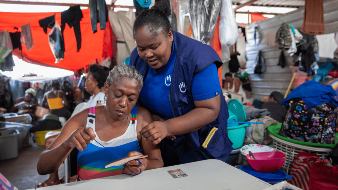 A seated woman holds a wooden object through which she threads a lace. A woman wearing a HI jacket leans over her. Behind them, we can make out tarpaulins, metal sheets, clothes drying and people sitting down.; }}