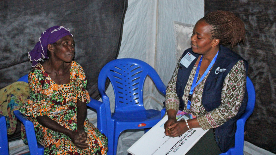 Two women are sitting on chairs in a tent. They are looking at each other and talking. The woman on the right is wearing a jacket with HI printed on it and is holding a notebook entitled “psychoeducation image box” on her lap.; }}