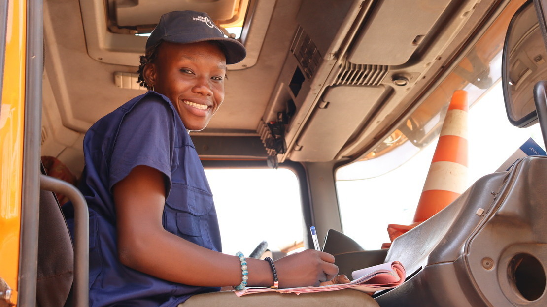 Une jeune femme noire est assise dans une cabine de camion. Elle tient un stylo et des papiers et regarde la caméra en contrebas en souriant.; }}