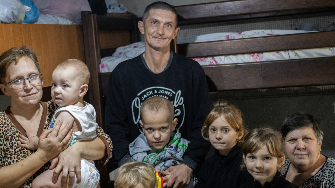 Une famille nombreuse est réunie dans une pièce avec des lits superposés en arrière-plan. Le père, au centre, arbore un léger sourire tandis que la mère, à gauche, tient un bébé dans ses bras. Autour d’eux, plusieurs enfants et leur grand-mère sont assis ou debout, regardant l’appareil photo avec des sourires légers. .; }}