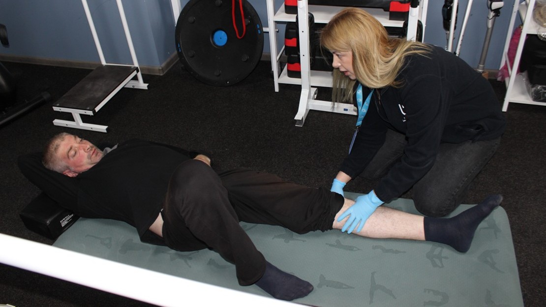 Une personne est allongée sur un tapis dans une salle de rééducation, avec une jambe pliée et l’autre allongée. Une autre personne, agenouillée à côté et portant des gants, maintient et manipule la jambe allongée au niveau du mollet. Du matériel de sport et de rééducation est visible en arrière-plan.