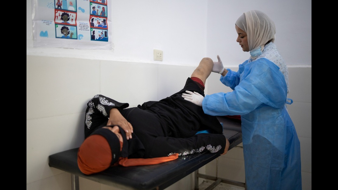   A person is lying on an examination table in a medical room. This person has had a leg amputated, and the stump is visible. The person is wearing dark clothing with shiny patterns on the sleeves, as well as a red cloth covering their head. They are lying on their back, with one hand resting near their face. Next to the table, another person is wearing medical attire consisting of a blue scrub top, gloves, and a mask positioned under the chin. This person is handling the stump with their hands. The room has white walls.   