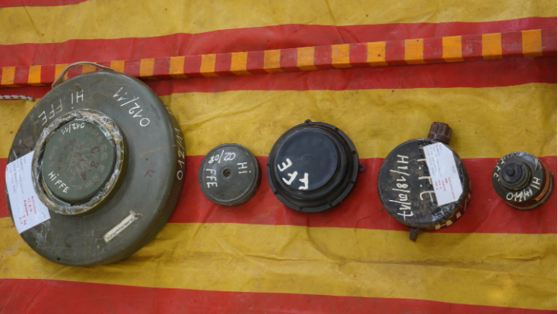 Five metal objects are arranged in a line on a yellow tarpaulin with wide red stripes. The objects are circular or cylindrical in shape and have worn and oxidised surfaces. They vary in size and bear hand-painted white inscriptions. The objects are displayed in an orderly fashion on the tarpaulin, in an outdoor or temporary setting, with natural lighting.