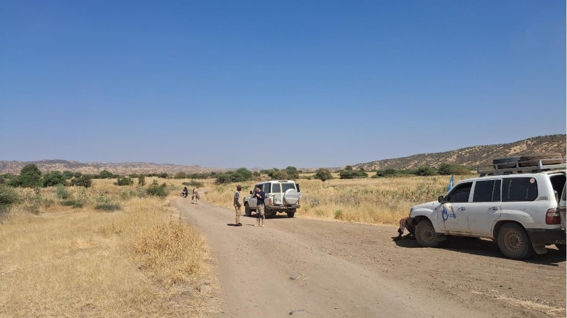 A team of four aid workers are standing and chatting on a path, near two HI vehicles.; }}