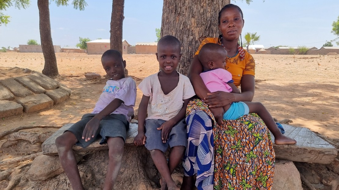 The image shows a woman and two children sitting side by side on a stone bench at the foot of a large tree. The woman is holding a child in her arms. The background shows a dry rural landscape with a few scattered trees and houses visible in the distance.; }}