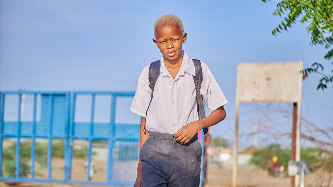 Portrait d'un adolescent en uniforme scolaire qui entre dans la cour d'une école en souriant. Derrière lui, on devine un portail en fer forgé bleu, un arbre et un grand ciel bleu sans nuage.; }}