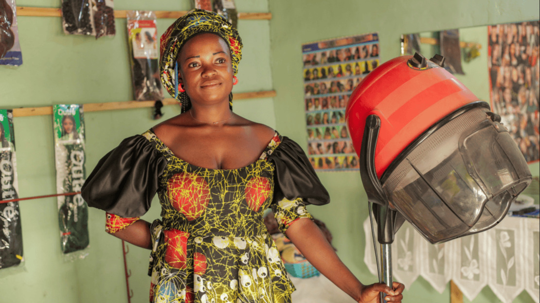 A smiling young woman is standing in a room whose walls are covered with posters of hairstyles and locks of hair. She is holding a hairdryer.; }}
