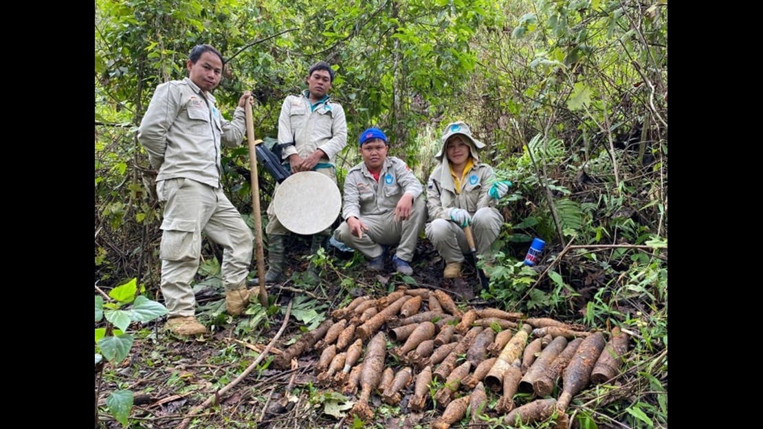 Quatre personnes se tiennent dans un environnement extérieur dense en végétation, entourées d’arbres, de buissons et de branches. Les personnes portent des vêtements de travail de couleur claire et des bottes. Certaines tiennent des outils, dont une pelle et un objet circulaire métallique. Au sol, devant les quatre personnes, une grande quantité d’objets métalliques allongés et rouillés est disposée en groupe. Les objets ont différentes tailles mais présentent des formes similaires. Une bombe aérosol est visible sur le sol à droite.; }}