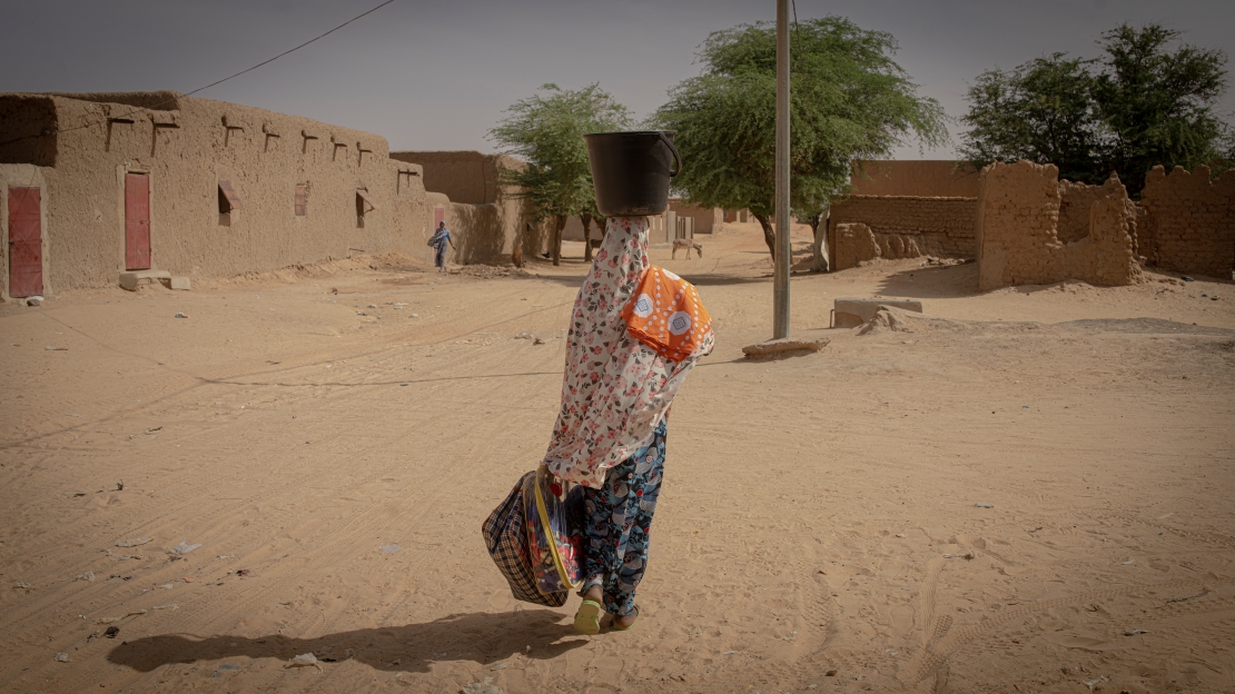 A woman walks with her back to the camera on a sandy path surrounded by mud houses and trees. She is wearing colorful clothes, with a black bucket on her head and a cloth over her shoulder. She is carrying two bags in her left arm.; }}