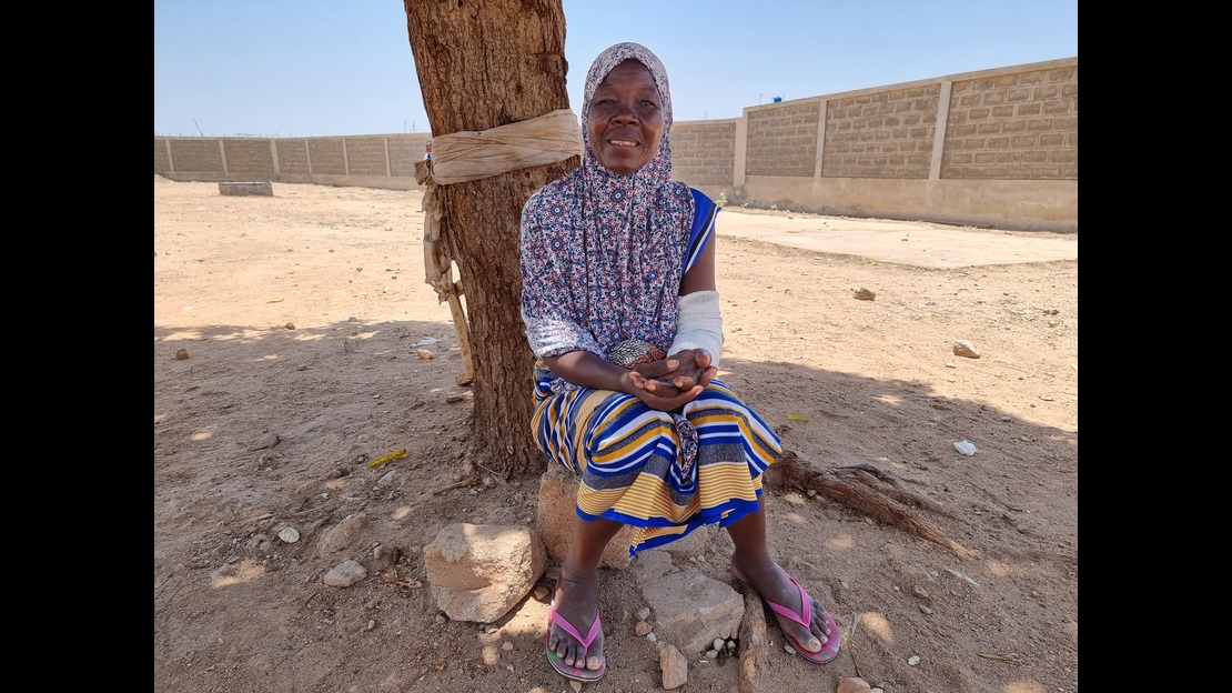 A woman is sitting on a rock at the foot of a tree in dry, sandy terrain. She is wearing a patterned top, colorful striped fabric, and sandals. A white bandage covers her right arm. In the background, a long brick wall surrounds the space under a clear sky.; }}