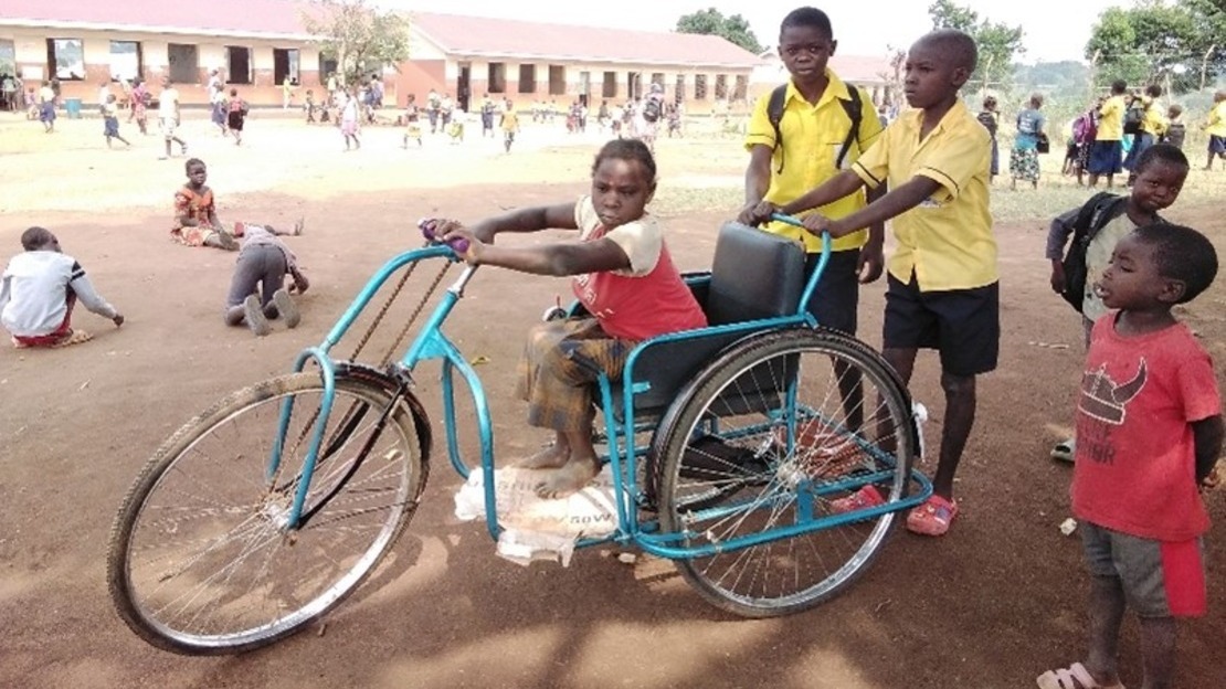 A young girl sits on a blue tricycle in a schoolyard. Behind her, two boys in school uniform push the tricycle.; }}