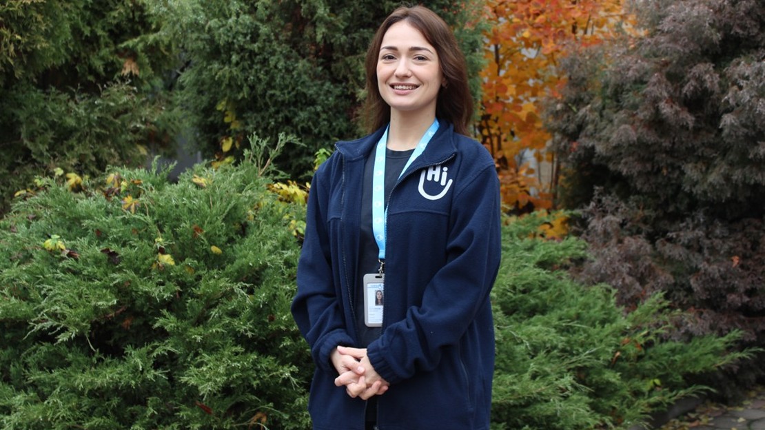A woman is standing outdoors, surrounded by dense vegetation consisting of green shrubs and trees with autumn leaves. She is wearing a badge around her neck and a dark blue top with a light-coloured logo on the chest, and is holding her hands clasped in front of her.; }}