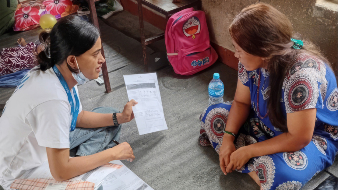 Two women are sitting in a room, facing each other; one is showing the other a sheet of paper whilst explaining something.; }}