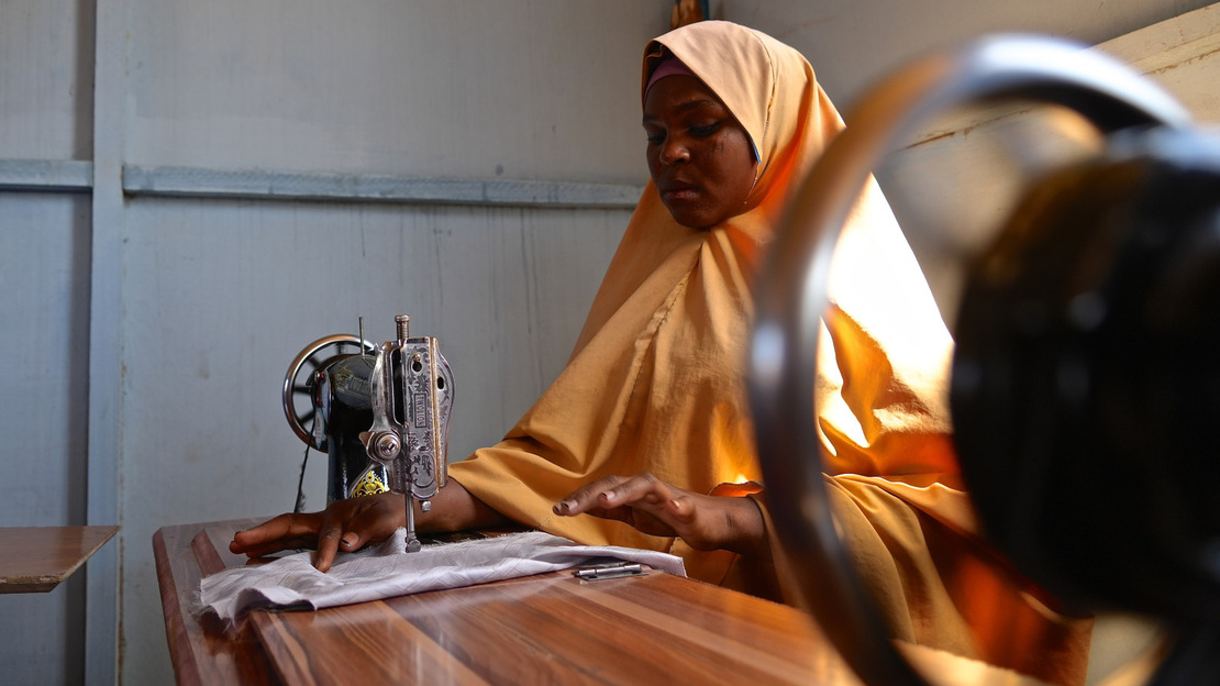 A young woman wearing a yellow veil sits at a table, sewing on her machine.; }}