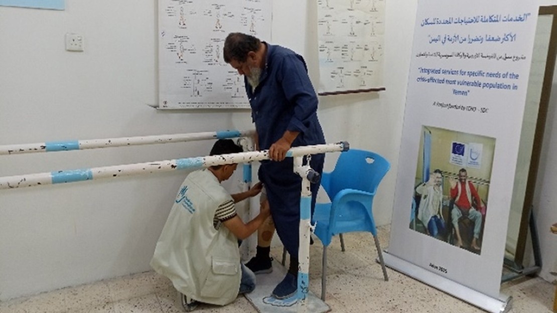 A person is standing between parallel rehabilitation bars in a room. Another person is fitting a prosthetic leg to the person’s lower limb. The room contains technical posters, a blue plastic chair, and an information panel on inclusion and rehabilitation.; }}