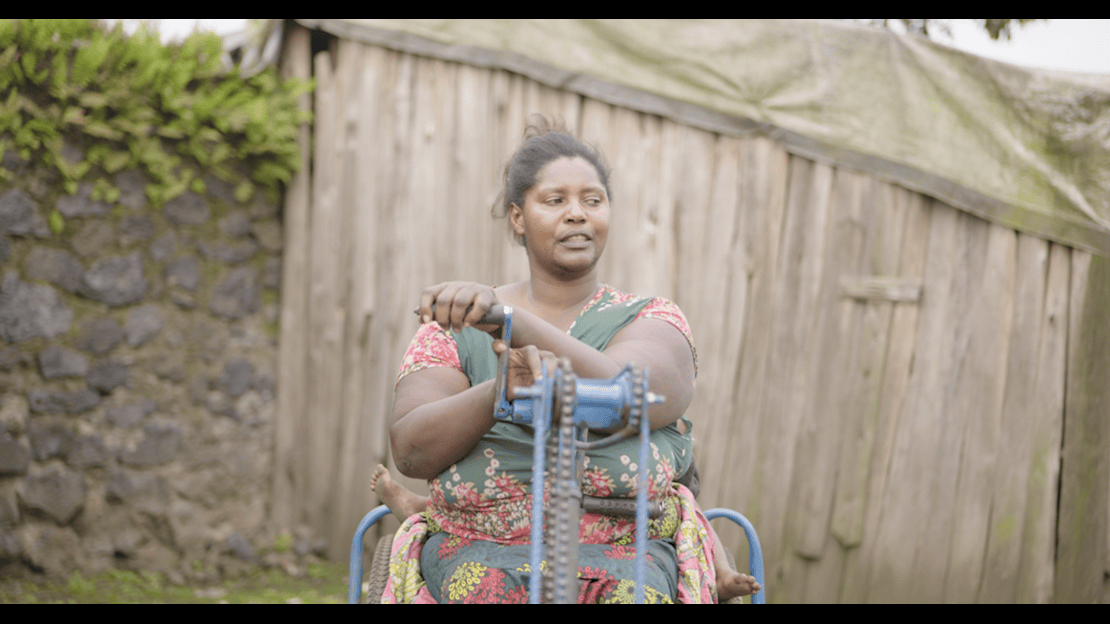Une personne est assise sur un tricycle à l’extérieur. Elle a posé ses deux mains sur un mécanisme manuel à manivelle et à chaîne placé à l’avant du tricycle. Un bébé est installé derrière elle dans le tricycle. La personne porte un vêtement sans manches à motifs et a les cheveux attachés. L’arrière-plan montre un bâtiment en bois aux planches verticales et un mur en pierre, dans un environnement rural avec de l’herbe au sol. 