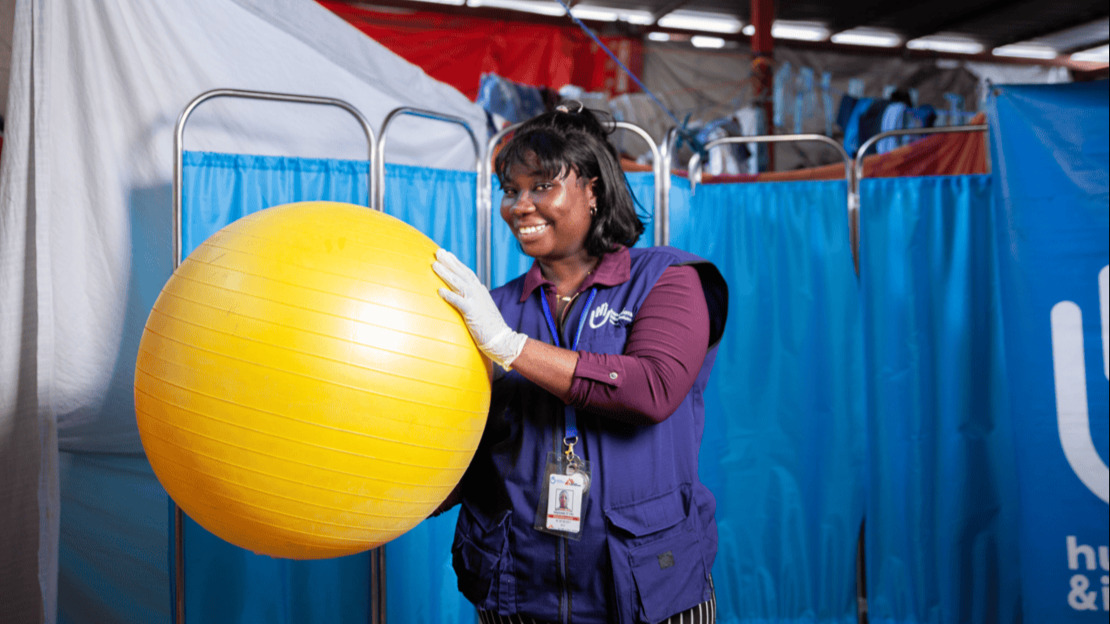 Portrait of a smiling woman wearing a latex glove and a HI jacket, holding a large yellow physiotherapy ball.; }}