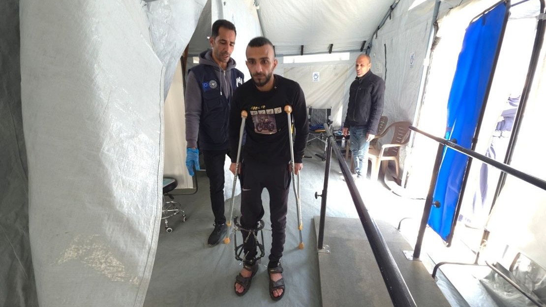 Inside a large, light-colored canvas structure, three people are standing in an area with a flat floor and a ramp equipped with metal handrails. In the foreground, a person is walking with two crutches, wearing a visible support device on one leg, and is dressed in dark clothing and sandals. Just behind them, another person stands nearby, wearing a dark jacket and blue gloves. In the background, a third person stands near plastic chairs and equipment arranged against the wall.; }}